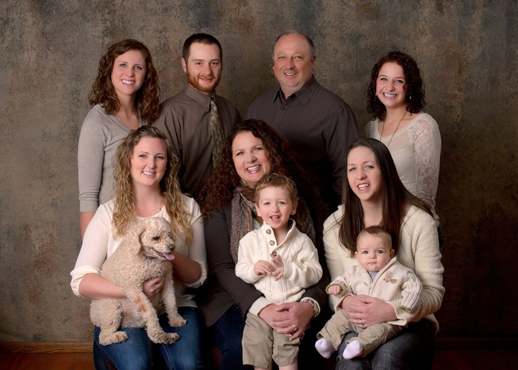 Multi-generational family portrait in a studio: adults, two young children, and a fluffy poodle, all smiling happily.